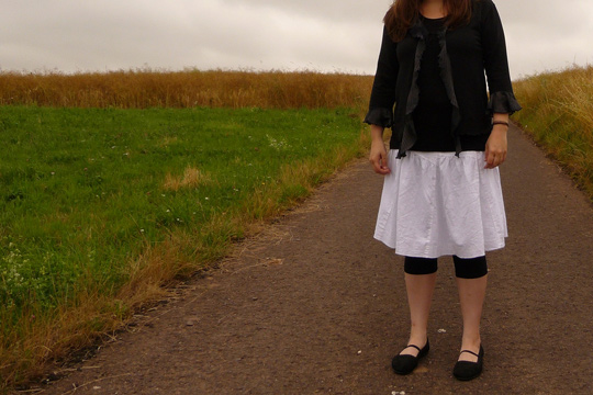 Mädchen mit weißem Rock und sonst schwarzer Kleidung steht auf einem Feldweg, der Kopf des Mädchens ist nicht zu sehen