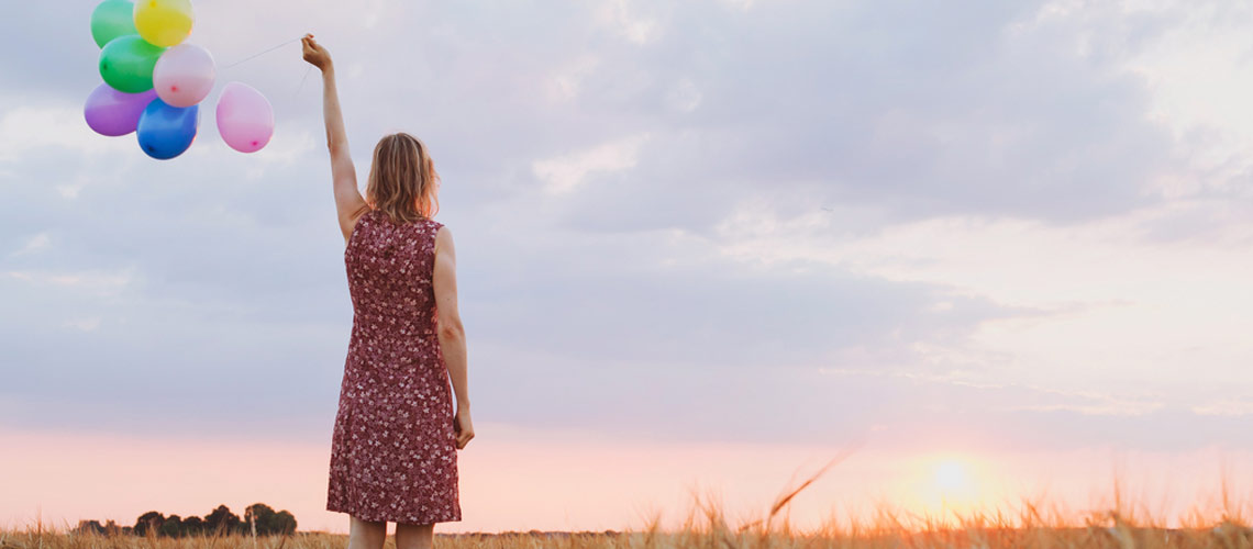 ine junge Frau steht auf einer Wiese, hält in der linken erhobenen Hand mehrere Luftballons in die Höhe, im Hintergrund der wolkenverhangene Himmel mit Sonnenuntergang